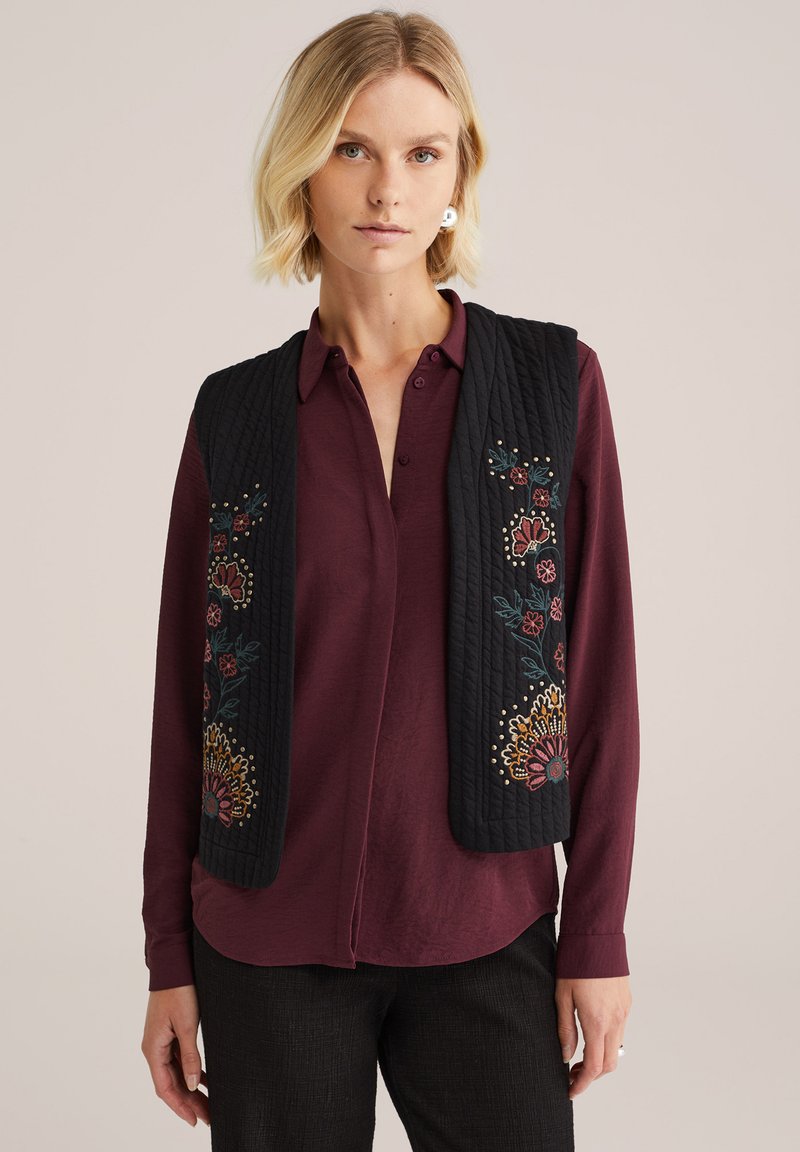 Zwarte gewatteerde gilet met bloemenborduurwerk in rood, geel en groen, gedragen over een bordeaux shirt. Heeft een recht model en een gedetailleerd patroon.