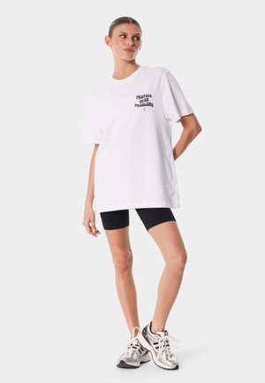 Młoda kobieta ubrana w białą oversize koszulkę z napisem „Protein Over Problems”, czarne szorty i srebrne buty do biegania, stoi z jedną ręką za plecami.