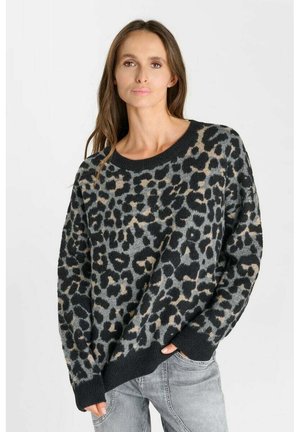 Vrouw draagt een losse grijs-zwarte trui met luipaardprint, met één hand in de zak van een lichtgrijze spijkerbroek, staand tegen een effen achtergrond.
