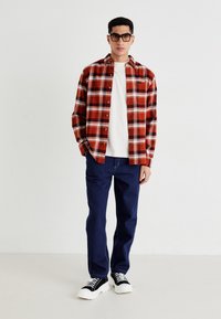 Un jeune homme se tenant debout, portant des lunettes noires, une chemise à carreaux rouges sur un t-shirt blanc, un jean bleu et des baskets noires avec des semelles blanches.