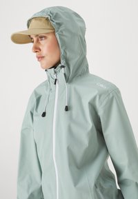 Lys grønn vanntett jakke med hette, frontlås og justerbare snorer. Kombinert med en beige caps. Glatt tekstur, minimal merking.