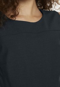 Svart tyg topp med en slät textur, rundad halsringning och subtil sömdetalj längs bröstet. Närbild av plagget.