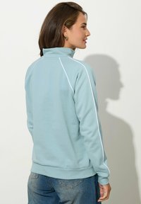 Maglione in pile azzurro chiaro con colletto alto, cuciture a contrasto bianche e polsini a costine, abbinato a jeans in denim. Texture liscia, vestibilità casual.