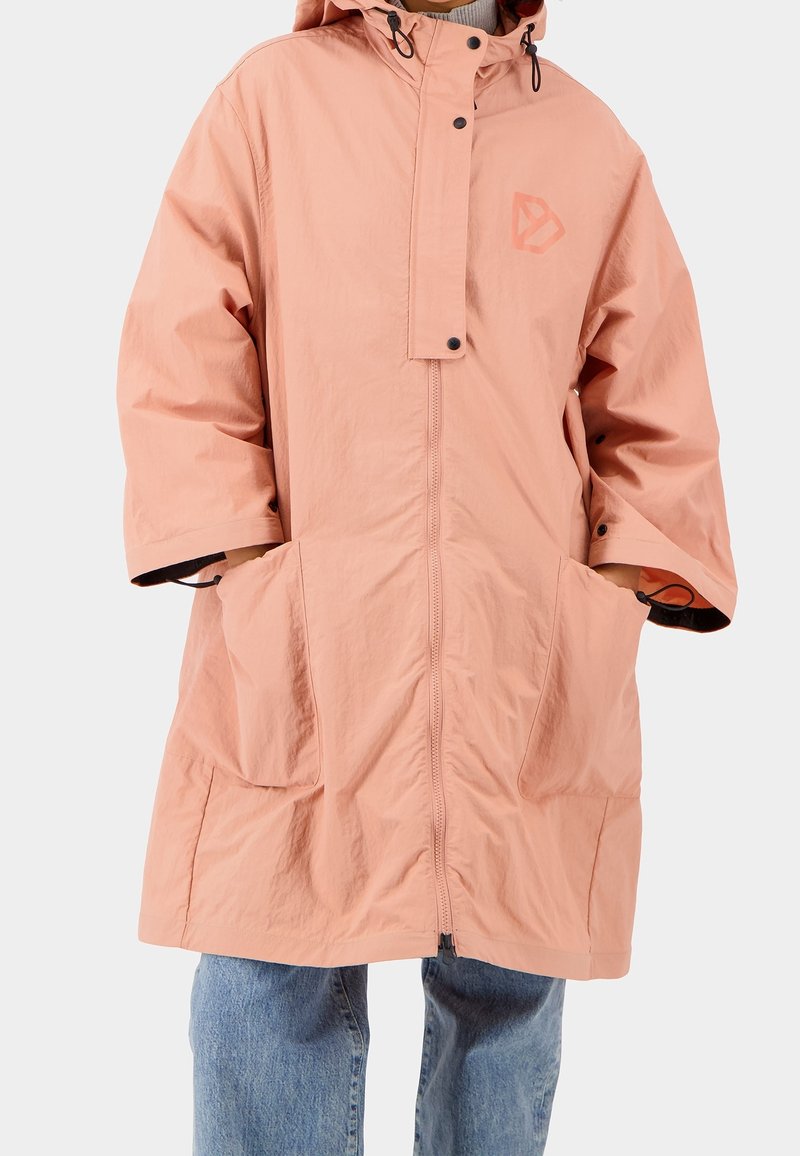 Personne portant un imperméable ample rose clair à capuche avec de grandes poches avant et une fermeture éclair frontale, par-dessus un jean bleu.