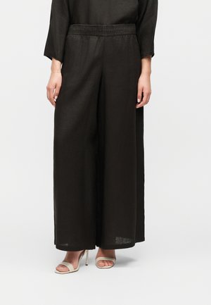 Femme portant un pantalon large noir à taille élastique et des sandales à talons hauts beiges, debout devant un fond uni.