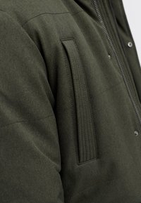 Tumeiroheline polsterdatud jope tekstuurse pinnaga, millel on vertikaalne lukuga tasku ja tasku kangal sooniline detail.