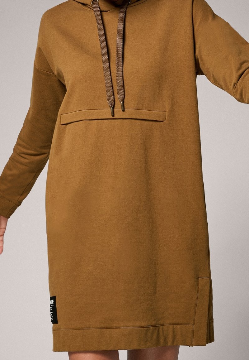 Robe à capuche marron à manches longues avec poche ventrale kangourou, cordons de serrage et étiquette noire près de l'ourlet, portée par une personne sur un fond uni.
