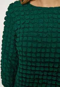 Zelený textúrovaný vrchný diel so mäkkým, nadýchaným povrchom s oblými zhlukmi, okrúhlym výstrihom a dlhými rukávmi.