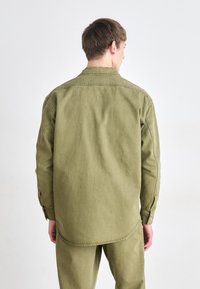 Khaki grønn langermet skjorte laget av slitesterkt stoff, med et ensfarget design, knappede mansjetter og rund kant bak.