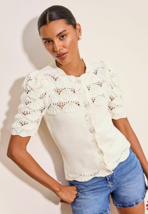 Femme aux cheveux foncés portant un haut court en dentelle blanche à manches courtes et un short en jean bleu, debout contre un mur clair uni.
