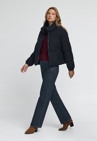 Mujer caminando vestida con una chaqueta acolchada negra, suéter burdeos, jeans oscuros y botas de tacón marrones, contra un fondo claro y sencillo.