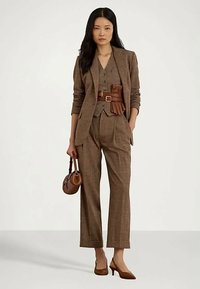 Nainen, jolla on päällään ruskea ruudullinen puku liivillä, leveä vyö, hänellä on ruskea käsilaukku ja ruskeat korkokengät. Hän seisoo yksinkertaista taustaa vasten.