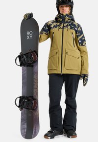 Deska snowboardowa ma ciemną górę z logo "ROXY", otoczoną czarnymi wiązaniami. Jeździec nosi złotą kurtkę z wzorzystymi rękawami i czarne spodnie.
