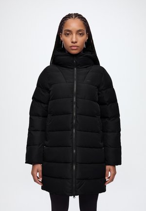 Une femme aux cheveux tressés porte un long manteau matelassé noir, à col montant, sur un fond blanc uni.