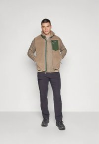 Chaleco de forro polar marrón con bolsillo verde en el pecho, cremallera frontal y tejido texturizado. Lleva pantalones oscuros y zapatos negros.