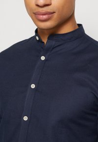 Camicia blu navy con colletto a band, realizzata in tessuto texturizzato, con chiusure a bottoni bianchi e dettagli di cucitura sottili.