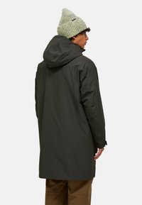 Parka impermeabilă de culoare verde închis, cu glugă, ajungând până la mijlocul coapsei. Mâneci mai lungi cu manșete reglabile. Purtați cu o căciulă tricotată de culoare verde deschis.