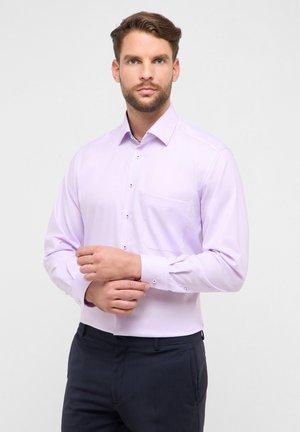 Camicia a maniche lunghe color lavanda chiaro con colletto, tasca frontale e chiusura con bottoni. Tessuto liscio e modello aderente. Indossata con pantaloni scuri.