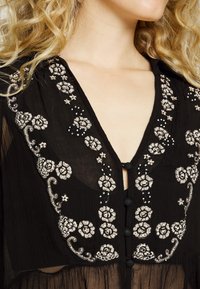 Blusa nera trasparente con intricati ricami floreali argentati, scollatura a V e dettagli con bottoni lungo la parte anteriore. Tessuto testurizzato.