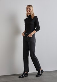 Top de cuello alto de manga larga negro, combinado con jeans de pierna recta oscuros de tiro alto y zapatos negros con cordones, de pie frente a un fondo neutro.