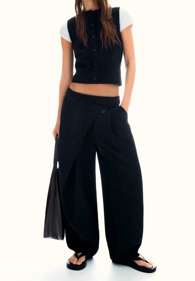 Femme portant un gilet noir sans manches boutonné sur un haut blanc à manches courtes, un pantalon large noir avec un rabat à l'avant, portant un sac noir, portant des sandales noires.