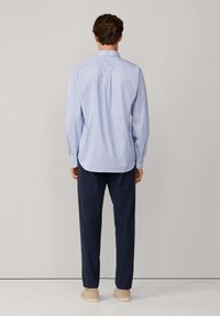Hombre de pie, de espaldas, vistiendo una camisa de mangalarga a rayas azul clara, pantalones azul oscuro y zapatos beige, contra un fondo neutro.