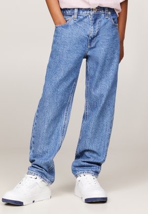 Barn iført løstsiddende lyseblå jeans og hvide sneakers, stående med hænderne i lommerne mod en ensfarvet baggrund.