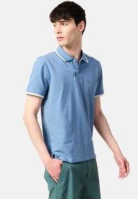 Vaalean sininen puuvillapolo-paita, jossa on valkoiset yksityiskohdat kauluksessa ja hihoissa. Paidassa on kolmen napin listaa ja kirjailtu logo rinnassa.