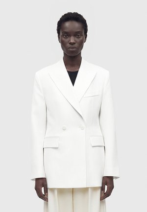 Femme portant un blazer blanc croisé sur un haut noir, debout devant un fond uni gris clair avec une expression neutre.