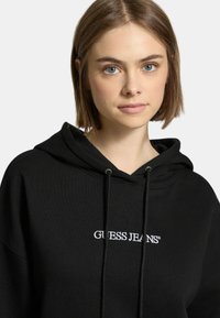 Felpa nera con cappuccio realizzata in tessuto morbido, con il logo ricamato bianco "GUESS JEANS" sul petto e dettagli con coulisse sul cappuccio.
