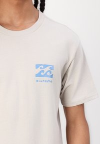 Bežinė medvilninė marškinė su stačiakampiu šviesiai mėlynu bangos grafiku ir "Billabong" tekstu kairėje krūtinės pusėje. Trumpomis rankovėmis, apvaliu iškirpimu.