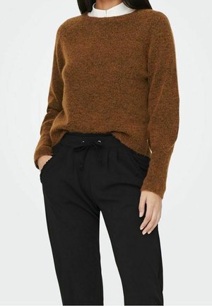 Femme portant un pull marron duveteux par-dessus une chemise blanche, associée à un pantalon noir à cordon, mains dans les poches, debout devant un fond uni.