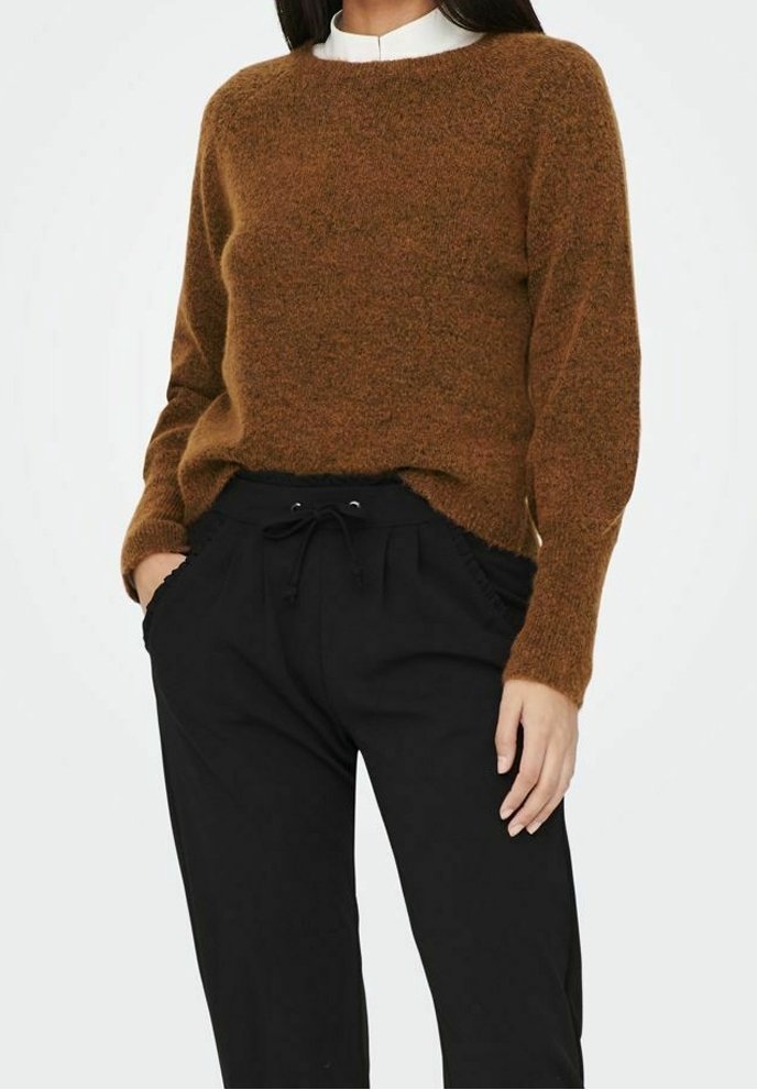 Femme portant un pull marron duveteux par-dessus une chemise blanche, associée à un pantalon noir à cordon, mains dans les poches, debout devant un fond uni.