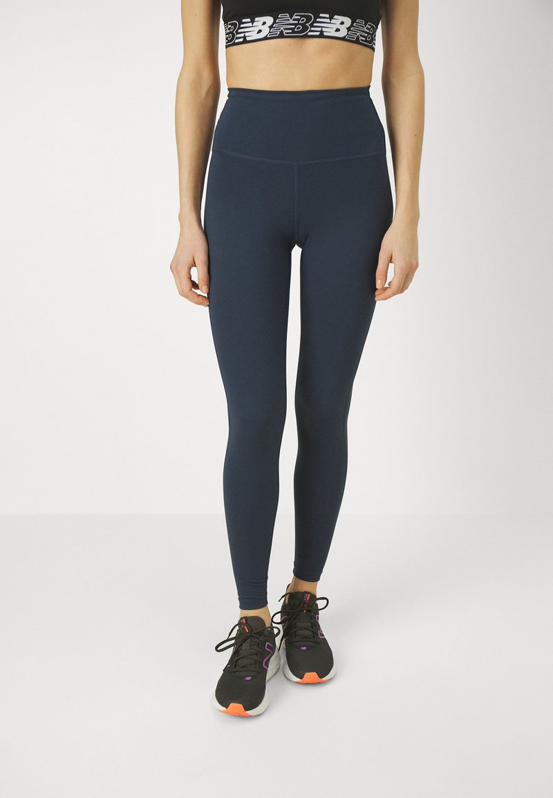 Femme portant des leggings taille haute navy, un brassière de sport noire avec un logo blanc, et des chaussures de course noires avec des accents orange et violet.