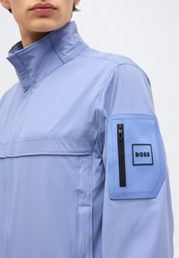 Άνδρας που φοράει ανοιχτό μπλε μπουφάν με ψηλό γιακά και τσέπη με φερμουάρ και ετικέτα "BOSS" στο αριστερό πάνω μανίκι.