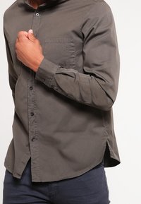 Camicia lunga a maniche verdi scuro con colletto a button-down, tasca sul petto a sinistra e orlo curva. Realizzata in un tessuto liscio con una consistenza morbida.