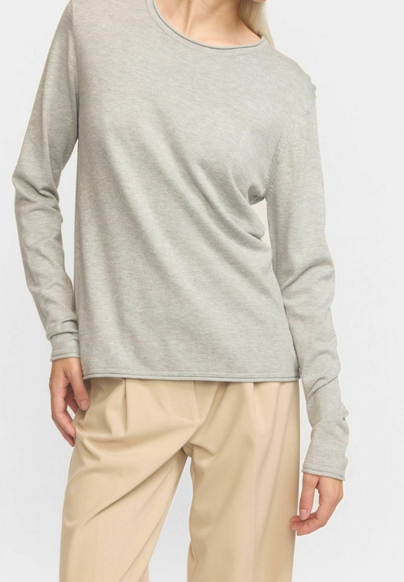 Personne portant une chemise gris clair à manches longues et un pantalon plissé beige, debout devant un fond blanc uni. Visage partiellement hors du cadre.