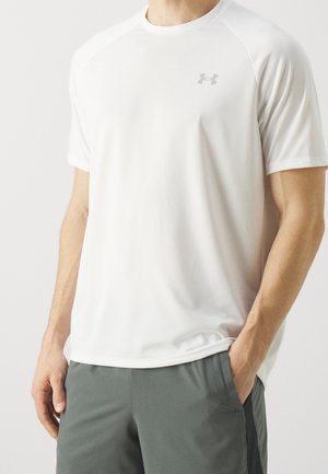 Homme portant un t-shirt de sport blanc uni à manches courtes avec un petit logo sur la poitrine et un short gris foncé.