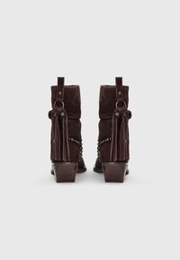 Botas de ante marrón oscuro hasta la mitad de la pierna con correas con flecos y bordes con tachuelas, tacones de madera apilados, mostradas desde la parte posterior sobre un fondo blanco.