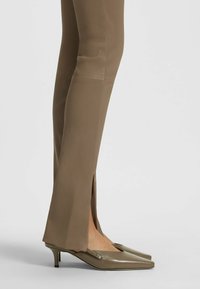Scarpe con tacco alto verde oliva con punta affusolata e chiusura con zip, abbinate a pantaloni marroni aderenti con spacchetti laterali all'orlo.