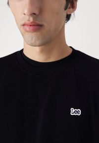 Jonge man draagt een zwart sweater met ronde hals en een klein wit "Lee"-logo op de linkerborst, neutrale achtergrond.