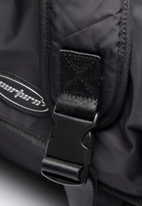 Black nylon strap with a plastic buckle, featuring textured surface and stitching details, attached to a bag with a logo.