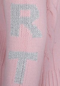 Manica di un maglione a maglia rosa chiaro con le lettere metalliche argento "R" e "T". La texture è morbida con un dettaglio a coste sul polsino.