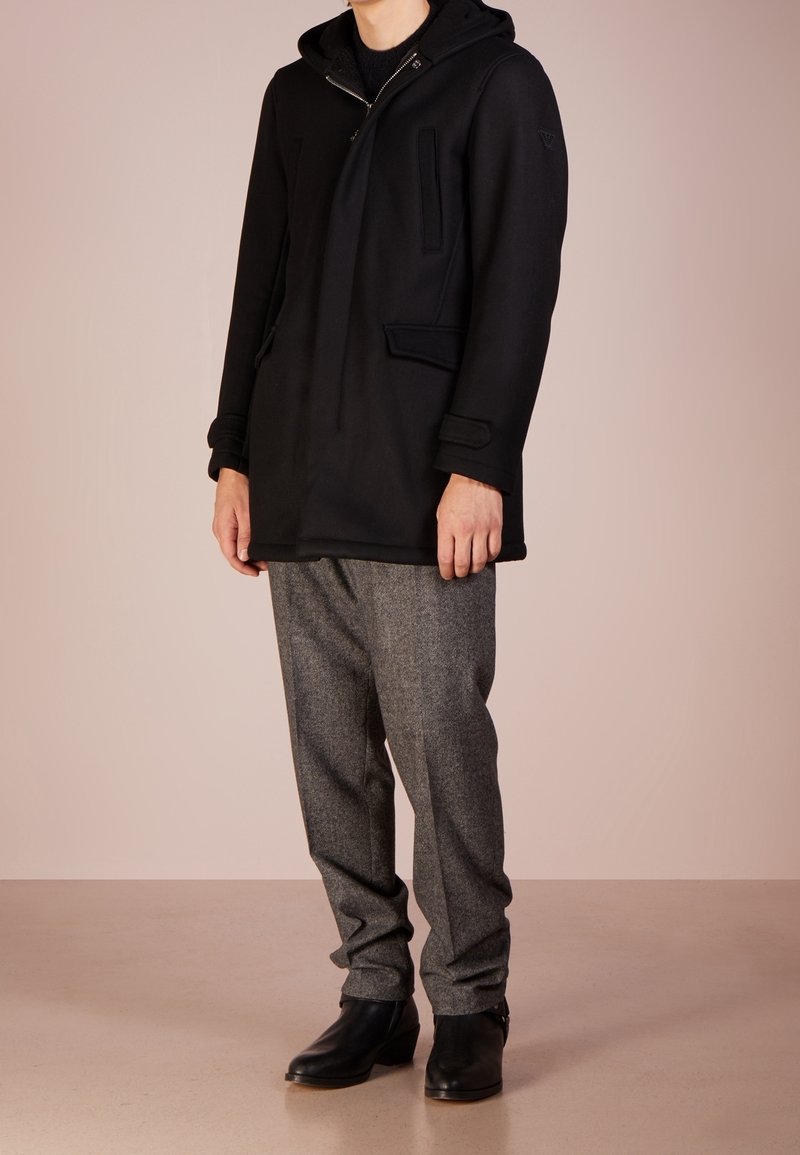 Cappotto esterno nero con cappuccio, caratterizzato da una cerniera frontale, due tasche laterali e spalle strutturate, abbinato a pantaloni grigi con texture e stivali neri.