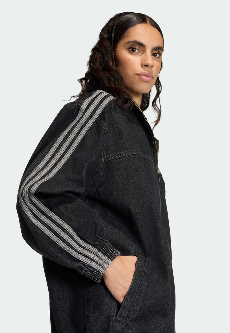 Black denim jacket with zipper, ribbed cuffs, and grey striped accents on sleeves. Model poses with hands in side pockets, showcasing fit.