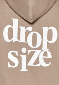 Beige Kapuzenpullover mit Kordelzug und großem weißen Text, der prominent auf der Vorderseite "drop size" zeigt. Weicher Stoff.
