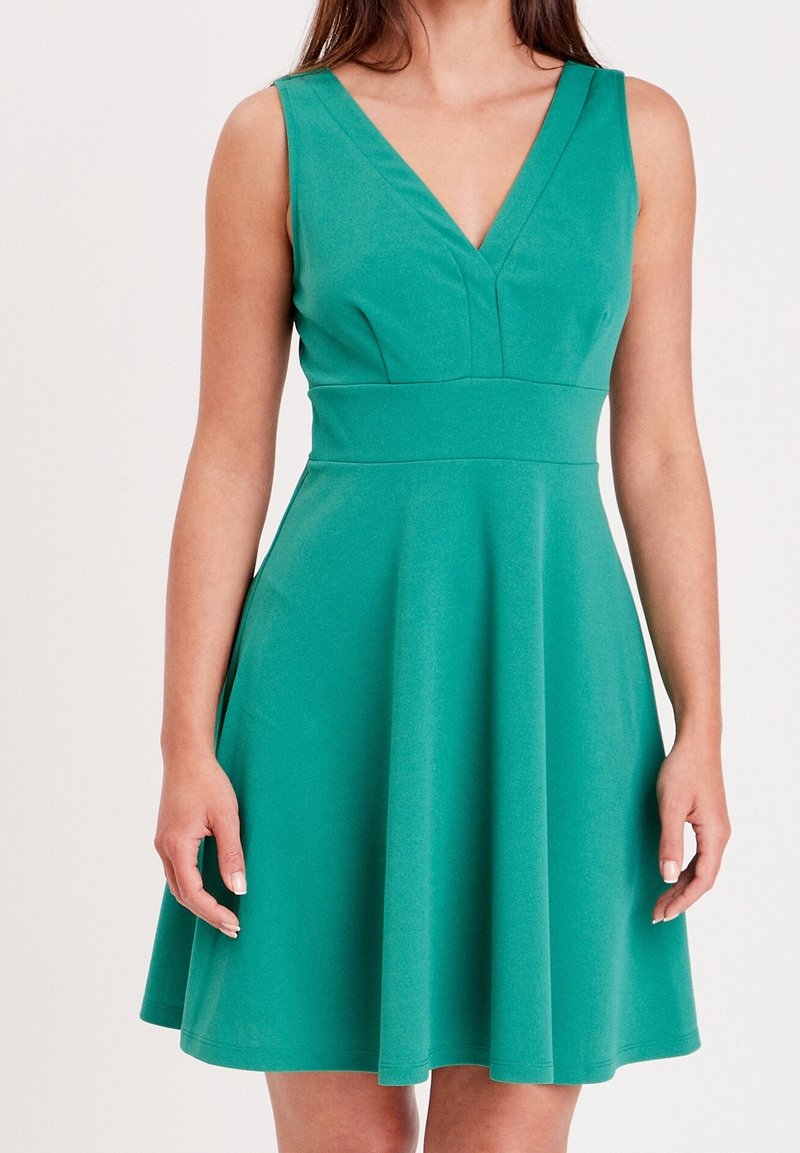 Femme portant une robe verte sans manches avec un décolleté en V et une taille cintrée, debout devant un fond clair uni.