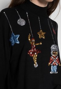 Pull en tricot noir avec des motifs colorés en sequins, comprenant des étoiles, un danseur et un soldat de jouet, avec un col montant et une finition texturée.