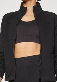Chaqueta negra de cremallera en tela suave, con cuello alto, logo de la marca sutil y diseño ajustado, combinada con un sujetador deportivo negro debajo.