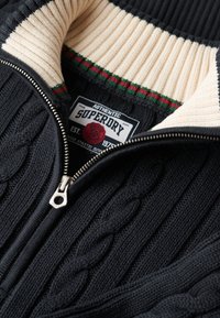 Schwarzer gestrickter Pullover mit cremefarbenem, geripptem Kragen, versehen mit einem Reißverschluss und grünen sowie roten Streifendetails. Das Etikett trägt die Marke "Superdry".